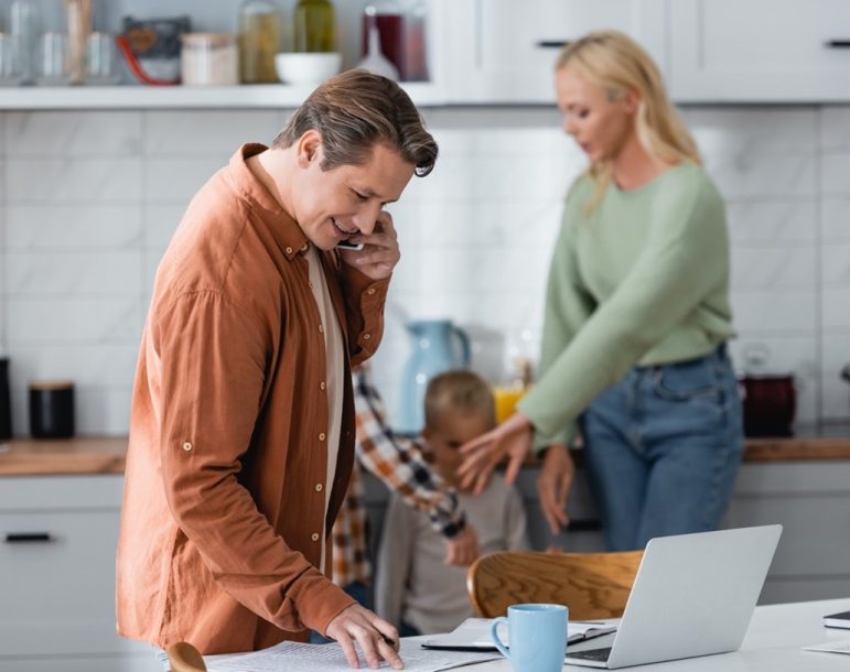 In the foreground, a man speaks on a cellphone while reviewing documents in front of a laptop at a kitchen table. In the background, a woman gives instruction to two young children.